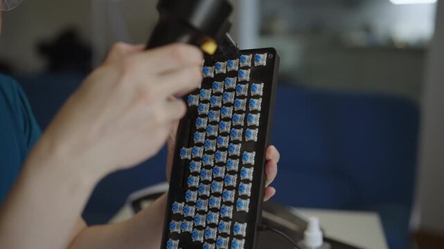 Close-up shot of person hands using handheld air duster to blow dust from blue switches of custom mechanical keyboard. Concept of detailed maintenance and care of computer hardware