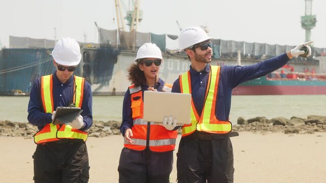 Team of industrial engineers in safety gear discuss project plans on laptop at port, with cranes and cargo ship in background.
