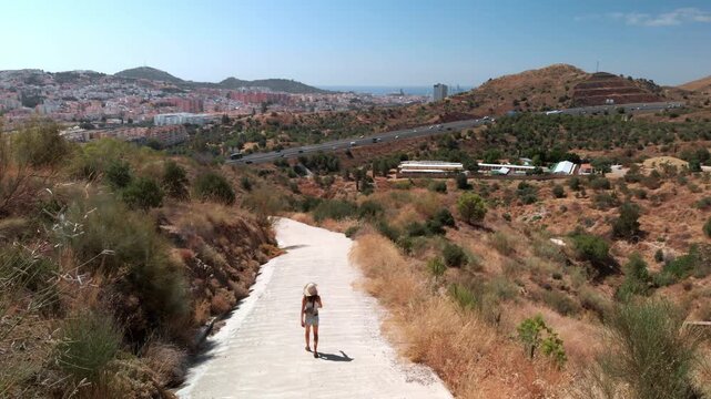 Woman walking down a scenic path on a dry hillside overlooking Malaga city and highway, exploring the urban landscape and coastal views, conveying a sense of journey and discovery.