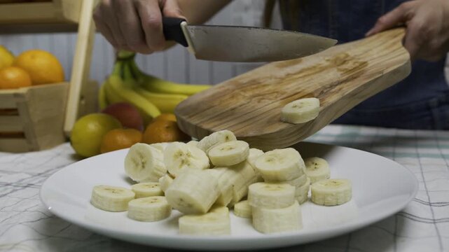 Slow motion closeup shot of woman using knife to push banana slices off cutting board and onto plate