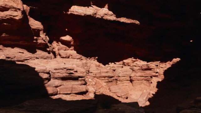People walk on a rocky surface inside a canyon. Sunlight creates shadows and highlights on the red rocks. The deep canyon walls rise steeply around them.