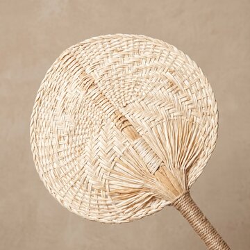 Round woven straw fan with a braided handle resting against a neutral textured light brown backdrop.