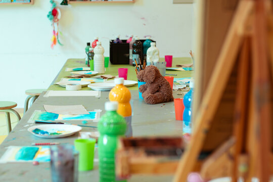 Children's art workshop featuring a messy table with colorful paint bottles, brushes, paper, and a teddy bear in a bright, creative environment