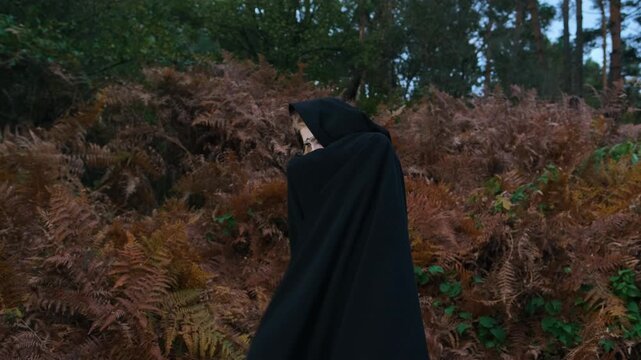 Hooded figure in matte black cloak raises sleeve across face, partly covering pale painted features with dark eye makeup, red lips, and forehead dots amid blurred autumn woodland foliage