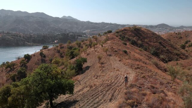 Woman hiking alone on dry hillside path overlooking Embalse del Limonero reservoir near Malaga, Spain, exploring arid landscape with distant city views, conveying solitude and peaceful outdoor adventu