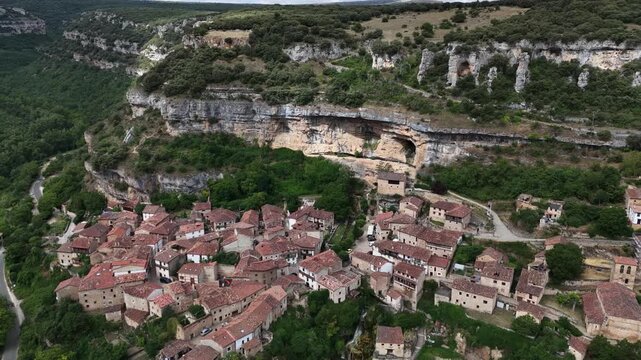  cliffside village scene in mountainous terrain with cobblestone streets and traditional stone houses with red-tiled roofs, aerial view. Risco los camellos, Cascada de Orbaneja del Castillo