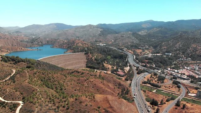 Aerial drone gliding over Embalse del Limonero reservoir dam in Malaga, Spain, showcasing vast blue water against arid mountainside, communicating vital water resource management and serene natural be