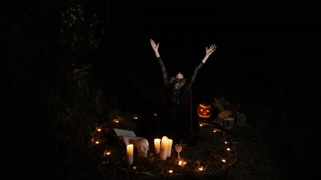 Kneeling woman with long blonde hair raises both arms and tilts head back inside rough circle of lit candles and tealights beside glowing pumpkin, suggesting dramatic ceremonial movement