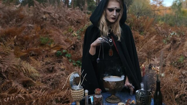 Woman dressed in black hooded cloak stands behind ritual table in woodland clearing, holding pendant above bronze cauldron releasing white vapor while gazing downward with somber expression and