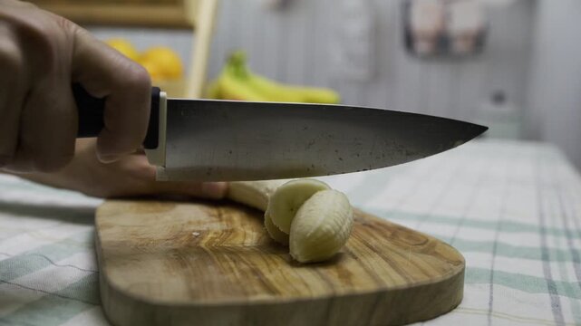 Closeup, slow motion shot of woman using knife to cut banana on cutting board in kitchen