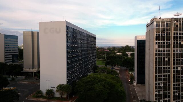 Aerial drone video shot moving past the large CGU office building. The scene overlooks streets, neighboring institutional structures like INSS, and lush green urban areas under a cloudy sky.