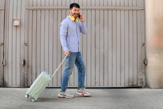 Smiling traveler pulling suitcase while talking on smartphone