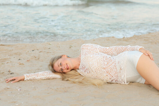 A young woman in a white lace outfit lies on the sandy shore near gentle sea waves with arms outstretched, peacefully resting as the tide softly reaches the beach