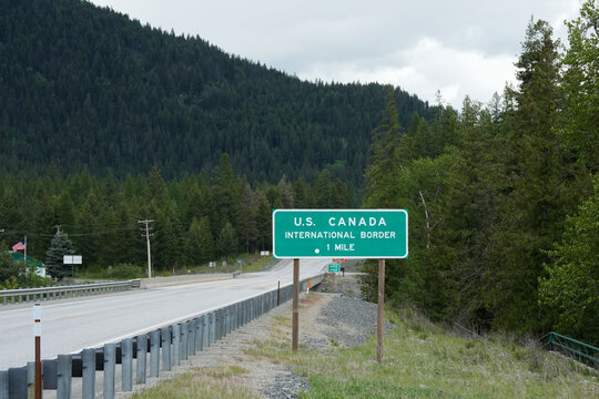 Green highway sign reading "U.S. Canada International Border" stands beside a two-lane road bordered by a metal guardrail, with forested mountains rising in the hazy distance under an overcast sky. 
