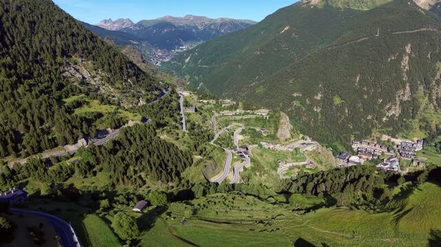Aerial view of a serpentine switchback road descending through dense pine forests toward the village of Canillo, Andorra, with Pyrenean peaks and a green valley beyond