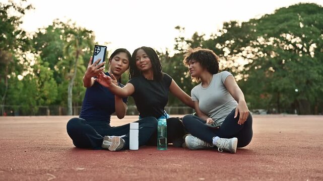 Three multicultural women sitting together on a sports court, taking selfies and laughing