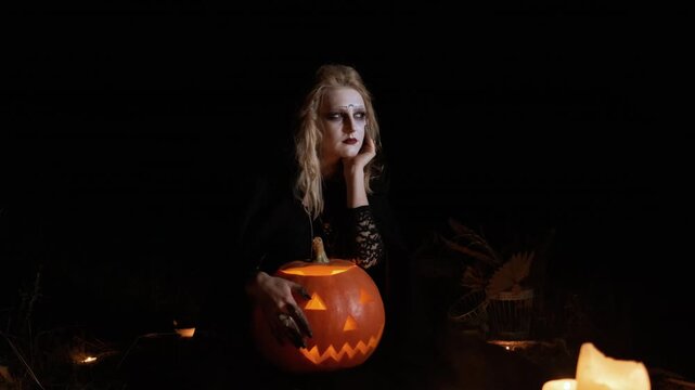Seated young woman in dark gothic costume cradles lit carved pumpkin while resting chin on one hand, gaze turned slightly off camera with pensive expression and moody candlelit atmosphere