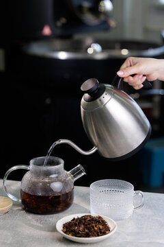 Pouring hot water from stainless kettle into glass teapot with loose cascara coffee grounds for brewing on roasting shop