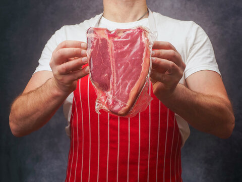 Masculine male butcher in white shirt and red and white classic stripe apron showing top quality T-bone steak in vacuum package for extra freshness of the product. Top price beef promotion.