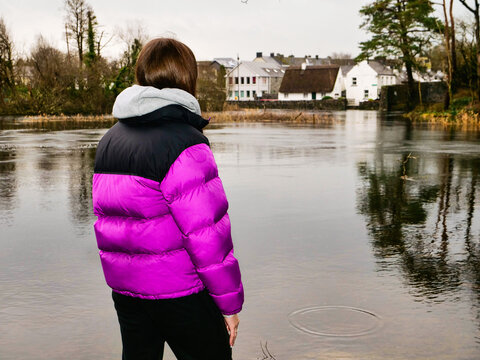 Young girl in purple puffy jacket is on a walk in an old park on a rainy day. Outdoor activity. She is looking at calm river and white old buildings in the background. Travel and tourism.