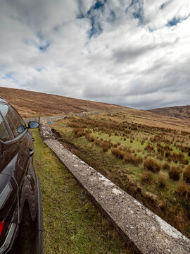 Black car parked beside old stone wall on rural road. Hills and grassy fields stretch into distance. Sky filled with scattered clouds. No animals or plants clearly identifiable