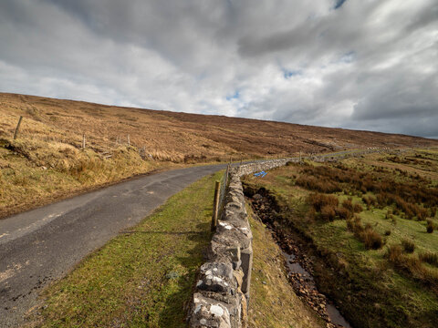 Road winds through open moorland under cloudy sky. Dry grasses line the path. Hills stretch into distance. No animals or specific plants visible