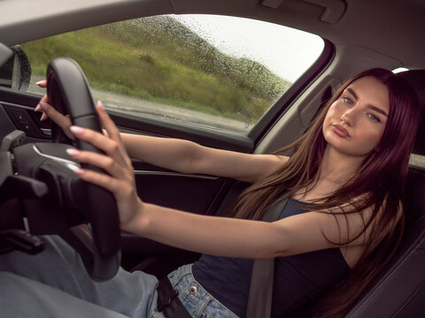 Young woman drives modern car. She looks ahead with calm focus. Hands rest on steering wheel. Car interior shows clean, sleek design. Outside view shows green landscape
