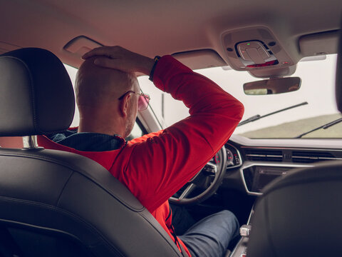 Man drives car, hands on steering wheel. Red jacket, bald head visible from back seat. Interior view shows dashboard and windshield. Daylight outside, road ahead. No animals or plants present
