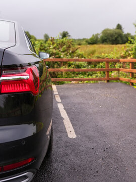 Black luxury car parked on asphalt near wooden fence. Greenery and field visible in background. Overcast sky above scene. No animals or plants clearly identifiable
