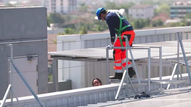 Two solar technicians working on rooftop photovoltaic installation, one drilling panel mounts while colleague checks system data on a tablet.