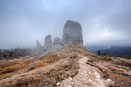 Cinque Torri emerges from low clouds beneath a dramatic overcast sky in the Dolomites. Atmospheric mountain landscape with fog, limestone towers and autumn colors