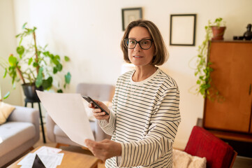 Obraz premium Middle aged woman standing with paper and smartphone