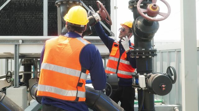 Two industrial technicians in safety helmets and vests adjusting a large valve on a heavy pipeline system at an industrial facility.