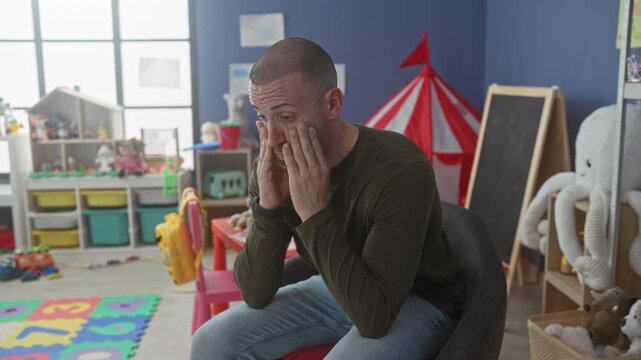 Man covering face with hands sitting on chair among toys and chalkboard in building kindergarten classroom; stress parenting.