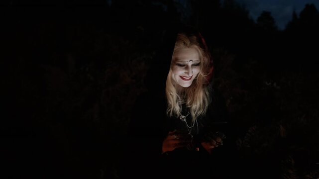 Pale-faced person in dark hooded cloak stands outdoors at night, holding small glowing object with open mouth, dark makeup, and dramatic shadows in eerie gothic forest scene, with person, hooded