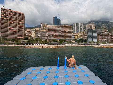 Woman sitting on floating plastic pontoon deck in Mediterranean Sea with Monaco skyline, modern luxury skyscrapers, Larvotto beach and mountains