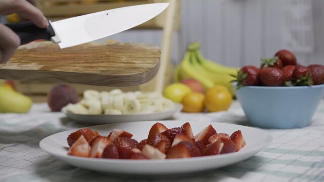 Person using knife to scrape cut strawberries onto white plate in kitchen - closeup