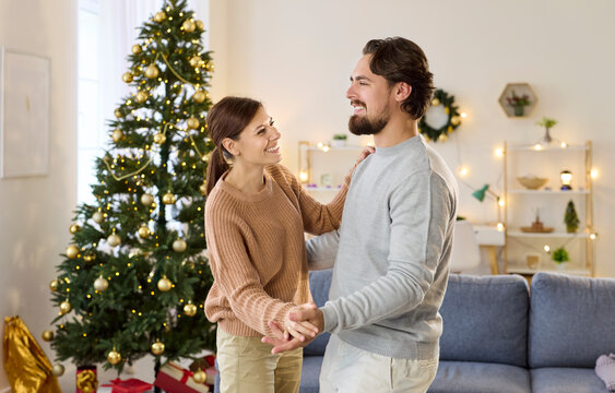 Couple dancing by Christmas tree at home. Twinkling lights brighten the living room as pair sway, enjoying music and winter holiday decor. Warm new year celebration, love, joy, and romance.