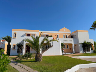 Fototapeta premium Mediterranean white and peach stucco resort villa with palm trees and lush green lawn under a bright clear blue sky on a sunny day