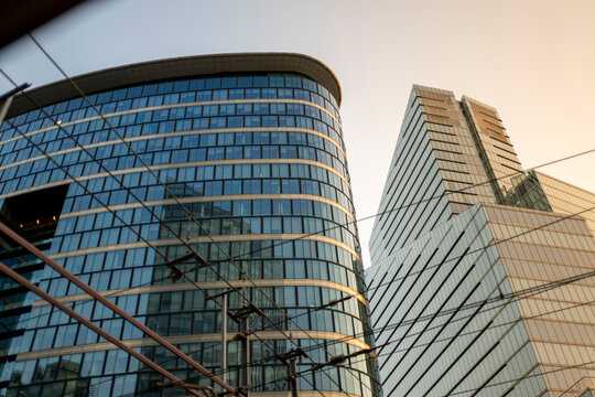 Curved glass office building beside a contemporary high rise tower viewed through railway catenary lines during golden hour in a modern business district