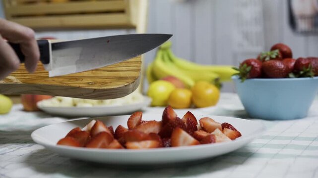 Knife being used to scrape cut up strawberries onto plate, closeup
