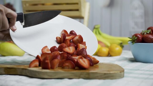 Person using knife to scrape strawberries from plate to cutting board, closeup shot