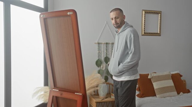 Man adjusts hoodie in front of full length mirror and bed in studio; quiet self reflection thoughtful.