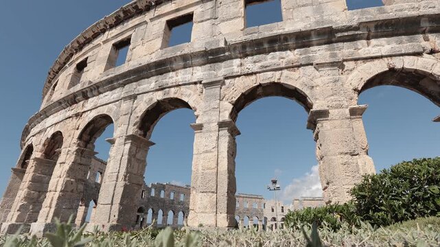 Ancient Roman arches of the Pula Arena rise above Mediterranean greenery under a clear blue sky. The cinematic shot captures the timeless beauty of one of Croatia's best-preserved amphitheatres