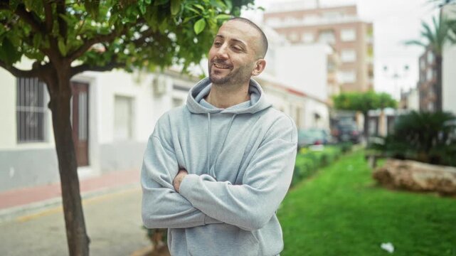 Man with visible face and arms crossed on street by building, wearing gray hoodie and smiling casually; contentment.