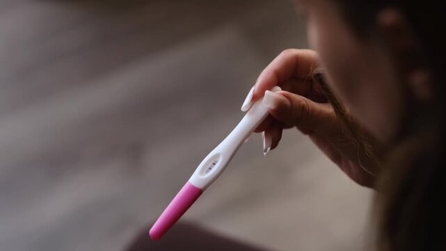 Woman examines pregnancy test in hand, displaying results with a focused expression, surrounded by soft lighting and a wooden floor