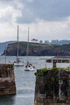 A vertical shot of two sailboats anchored in the calm waters of Candas harbor framing the green cliffs and industrial gas storage tanks in Asturias, Spain.