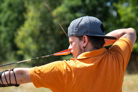 Bearded archer with cap and orange shirt aimed to his target, waiting with calm mind for the perfect time to release the arrow from the bow.