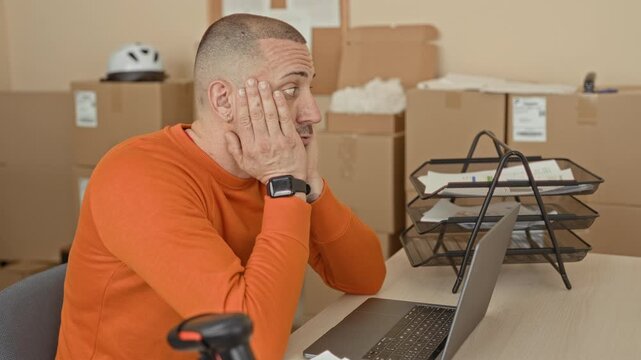 Man at laptop holding cardboard box rubbing temple while checking shipping labels and typing on laptop amid stacked parcels in building; work stress.