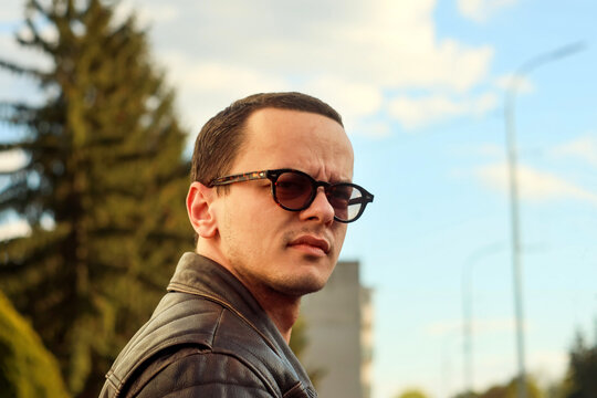 A portrait of a young man wearing stylish sunglasses and a leather jacket outdoors against a bright sky and trees background, illustrating confidence, fashion, and urban lifestyle